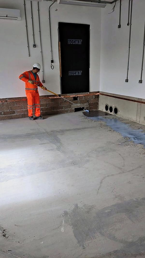 Operative applying a floor coating beside exposed brickwork and services