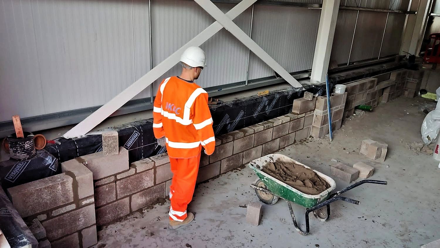 PKBC bricklayer laying block courses on a low wall inside a steel-framed building, mortar barrow alongside