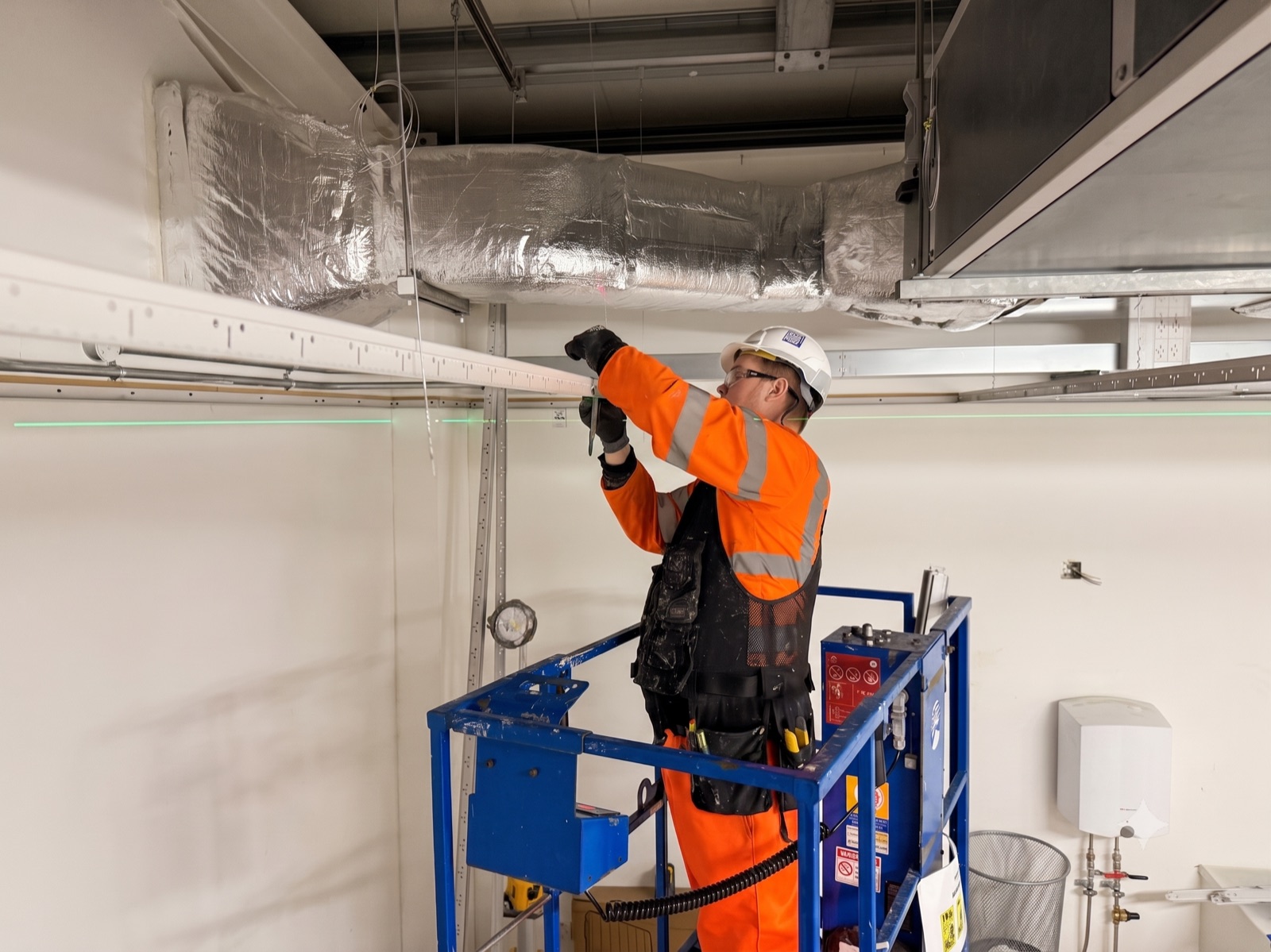 PKBC operative installing suspended-ceiling services from a scissor lift inside the Gremista substation building