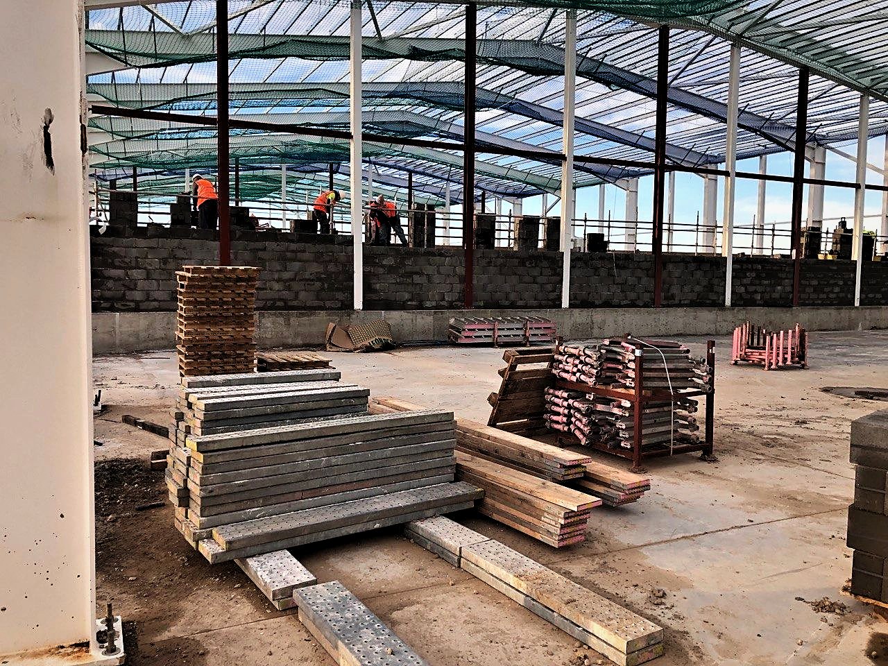 Bricklayers building a perimeter blockwork wall inside a netted steel-framed building