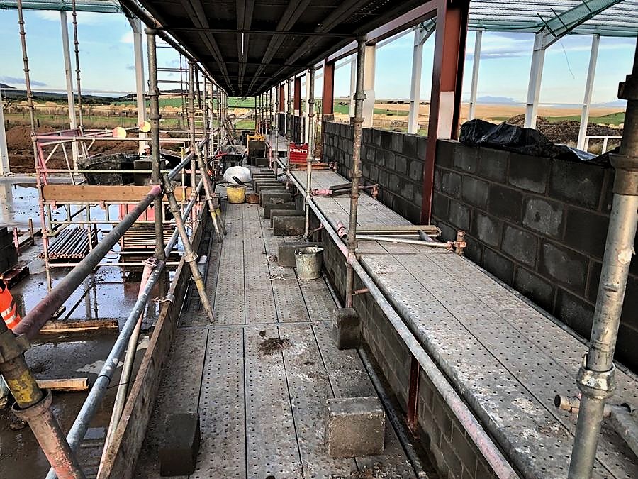 Scaffold access walkway along blockwork walls on a steel-framed building under construction, with open farmland beyond